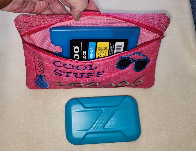 K&ouml;nnte beinhalten: Rosa isolierte K&uuml;hltasche mit dem gestickten Text "COOL STUFF" und Sonnenbrille. Die Tasche enth&auml;lt einen blauen K&uuml;hlakku. Ein zweiter blauer K&uuml;hlakku befindet sich unter der Tasche. Die Tasche ist ca. 10,8 x 21 cm gro&szlig;.