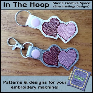 May include: Two keychains with a silver and white base, each featuring a pair of overlapping hearts in pink and red embroidery. The keychains are on a wooden surface. The text "In The Hoop by Sher" is visible in the lower right corner.