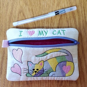 May include: A white zippered pouch with a purple zipper and a pink heart embroidered on the front. The pouch has the text "I love my cat" embroidered on the front. The pouch also has a colourful embroidered cat on the front.