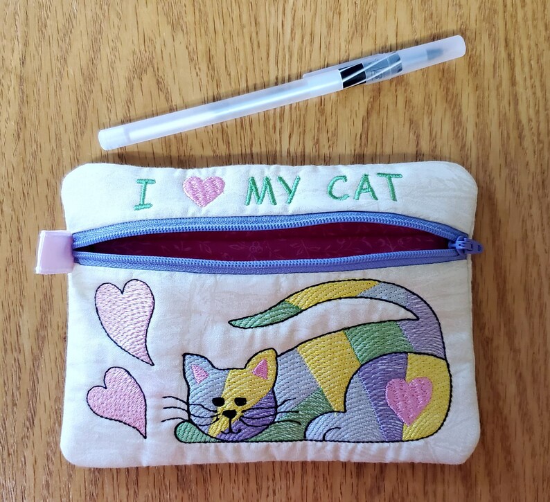 May include: A white zippered pouch with a purple zipper and a pink heart embroidered on the front. The pouch has the text "I love my cat" embroidered on the front. The pouch also has a colourful embroidered cat on the front.