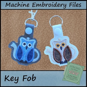 May include: Two machine embroidery key fobs shaped like owls with teapot features. One is dark blue with a silver lobster clasp, the other is white and brown with a silver split ring. Both have light blue plastic snaps and embroidered details.