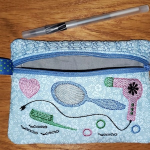 May include: A blue zippered pouch with a floral pattern and embroidered images of a hairdryer, a comb, a mirror, and a heart. The pouch is ideal for storing small items like hair ties, bobby pins, and other hair accessories.