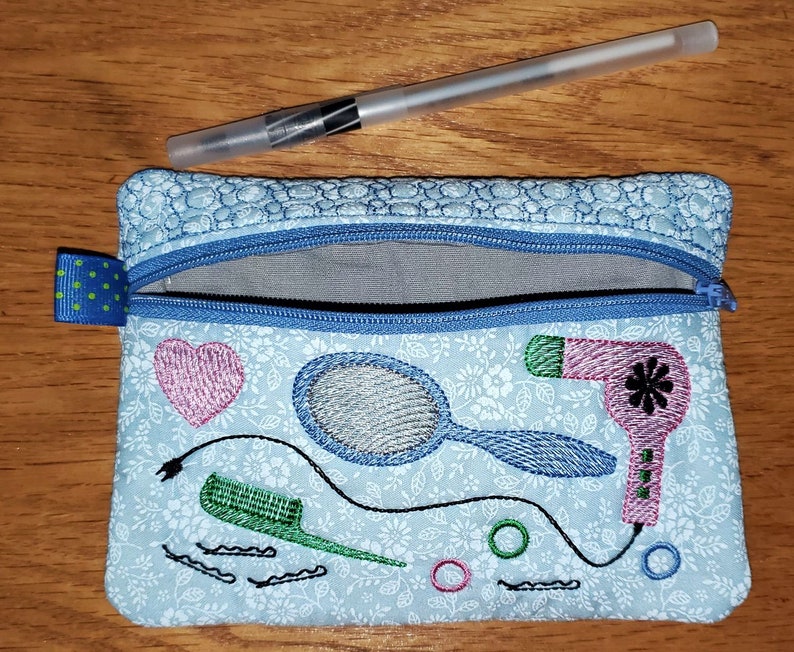 May include: A blue zippered pouch with a floral pattern and embroidered images of a hairdryer, a comb, a mirror, and a heart. The pouch is ideal for storing small items like hair ties, bobby pins, and other hair accessories.