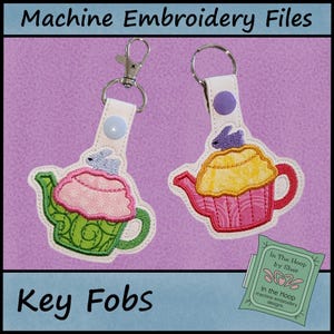 May include: Two key fobs with a teapot design. One is green and pink, the other is pink and yellow. Each has a metal clasp and a button closure. The text "Machine Embroidery Files" and "Key Fobs" are visible.
