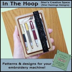 May include: A beige leather pen holder with a black elastic band. The pen holder has an embroidered design of five pens on a woodgrain background. The text "In The Hoop by Sher" is visible on a green tag.