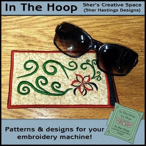 May include: A close-up of a small, rectangular, embroidered piece of fabric with a green floral design and a red flower in the center. The fabric is on a wooden surface with a pair of sunglasses on top of it. The fabric has a red and white border. The text "In The Hoop by Sher" is visible in the lower right corner of the image.