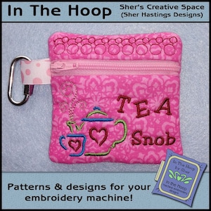 May include: Pink fabric pouch with a zipper closure and a metal clasp. The pouch has an embroidered design of a teapot and teacup with the words "TEA Snob" and a decorative pattern. The pouch is labeled "In The Hoop by Sher" and "In the Hoop machine embroidery designs".