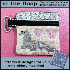May include: A small, beige zippered pouch with a gray seal and two pink hearts embroidered on it. The pouch has a black zipper and a black and white clip. The pouch is on a pink background. "In The Hoop by Sher" and "In the Hoop machine embroidery designs" are printed on a blue and green label.