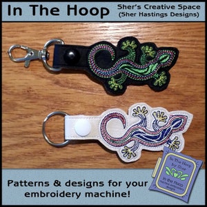 May include: Two embroidered keychains featuring a green and black lizard design and a blue and white lizard design. The keychains are on a wooden surface. The text "In The Hoop by Sher" is visible in the lower right corner of the image.