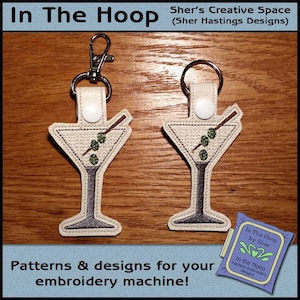 May include: Two embroidered keychains featuring a martini glass with olives and a sprig. The keychains are beige with a white button and a silver metal key ring. The text "In The Hoop by Sher" is visible in the lower right corner.