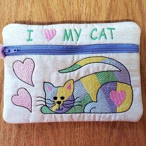 May include: A white zippered pouch with the text "I love my cat" embroidered on the front. The pouch features a colourful embroidered cat with pink hearts.