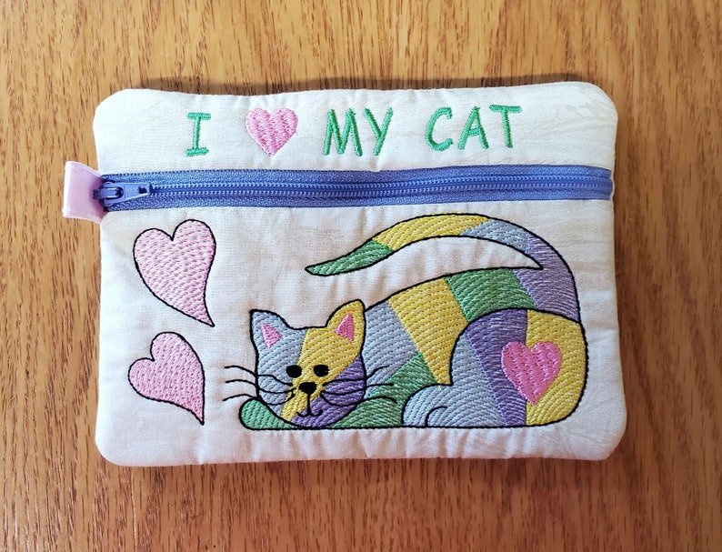 May include: A white zippered pouch with the text "I love my cat" embroidered on the front. The pouch features a colourful embroidered cat with pink hearts.
