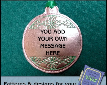 May include: A pink and green embroidered ornament with the text "YOU ADD YOUR OWN MESSAGE HERE". The ornament is on a green background. The text "In The Hoop by Sher" is visible in the lower right corner of the image.