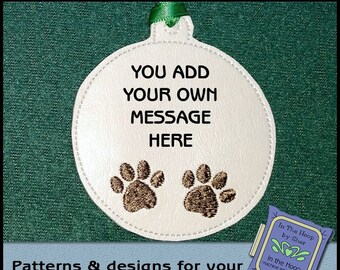 May include: White, round ornament with a green ribbon, featuring the text "YOU ADD YOUR OWN MESSAGE HERE" and two embroidered paw prints. The ornament is designed for machine embroidery. The image also includes the text "Patterns & designs for your embroidery machine!"