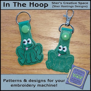 May include: Two green frog keychains with embroidered details. The keychains are attached to silver key rings. The keychains are on a wooden surface. The text "In The Hoop by Sher" is visible in the lower right corner of the image.