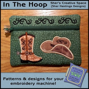 May include: A green zippered pouch with a cowboy boot and a cowboy hat embroidered on it. The pouch is on a wooden surface. The text "In The Hoop by Sher" is visible on a small tag.