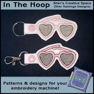 Puede incluir: Dos llaveros blancos con gafas de sol de corazón rosa bordadas en ellos. Los llaveros están sobre un fondo negro. El texto "In The Hoop by Sher" es visible en la esquina inferior derecha.