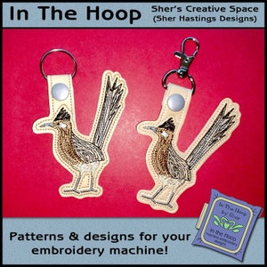 May include: Two embroidered roadrunner keychains with brown, white, and gray stitching. The keychains are attached to brown leather straps with silver key rings. The background is a bright red.