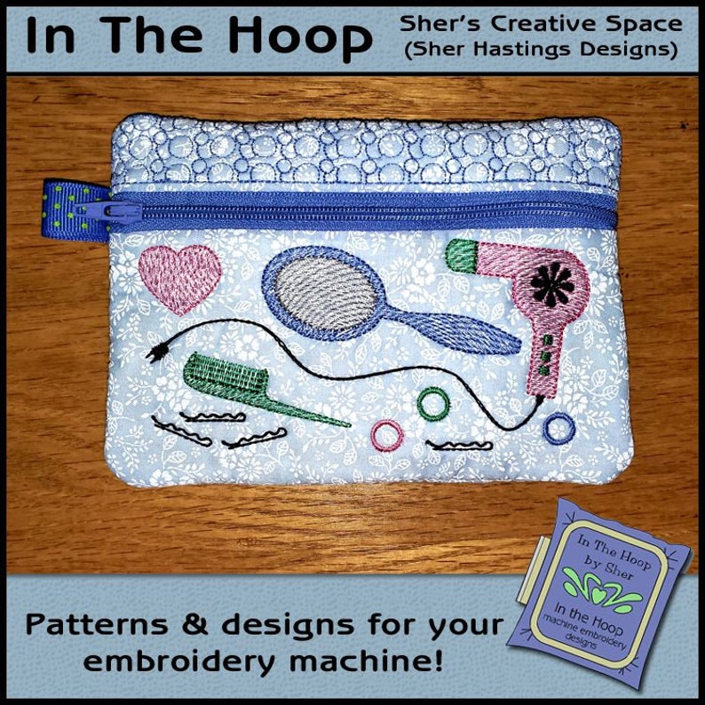 May include: A blue zippered pouch with a white background and a floral pattern. The pouch features embroidered images of a hairdryer, a mirror, hair clips, and a heart. The text "In The Hoop by Sher" is visible on a small tag.