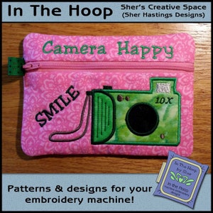 May include: Pink zippered pouch with an embroidered green camera and the text "Camera Happy" and "Smile" on it. The pouch is on a wooden surface.