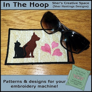 May include: A beige fabric coaster with a black silhouette of a cat and dog, pink hearts, and the text "In The Hoop by Sher" in a green box. The coaster is on a wooden surface with a pair of sunglasses on top.