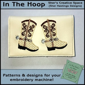 Könnte beinhalten: Eine weiße Lederbörse mit zwei gestickten Cowboystiefeln. Die Stiefel sind braun mit einem beigefarbenen Schaft und haben einen braunen Gürtel und Sporen. Der Text "In The Hoop by Sher" befindet sich auf einem grün-rosa Etikett in der unteren rechten Ecke.