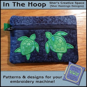 May include: A blue zippered pouch with two green embroidered sea turtles. The pouch is on a wooden surface. The text "In The Hoop by Sher" is on a white tag.
