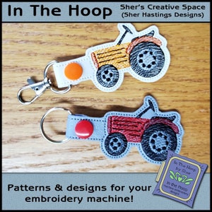 May include: Two embroidered tractor keychains, one red and one yellow, with a white background and a silver key ring. The keychains are on a wooden surface. The text "In The Hoop by Sher" is visible in the lower right corner of the image.