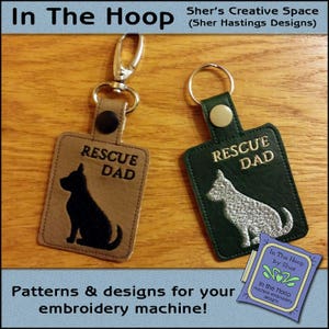 Könnte beinhalten: Zwei Leder-Schlüsselanhänger mit gestickten Hundesilhouetten und dem Text "Rescue Dad". Die Schlüsselanhänger sind braun und grün mit silbernen Glitzerakzenten.