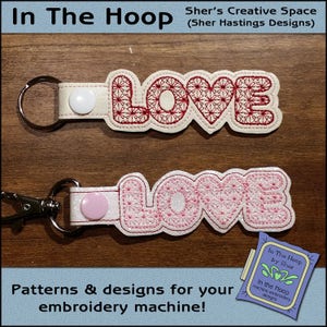 May include: Two white keychains with the word "LOVE" embroidered in red and pink thread. The keychains are on a wooden surface. The text "Sher's Creative Space (Sher Hastings Designs)" is at the top of the image. The text "In The Hoop" is at the bottom of the image.