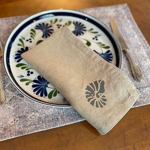 May include: A beige linen napkin with a dark gray nautilus shell design. The napkin is folded and placed on a blue and white patterned plate.