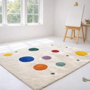 Pode incluir: Um tapete retangular branco sujo com grandes bolinhas coloridas em amarelo, azul, laranja, verde, roxo e vermelho. O tapete está colocado sobre um chão branco numa sala com uma janela, um cavalete e uma mesa pequena.
