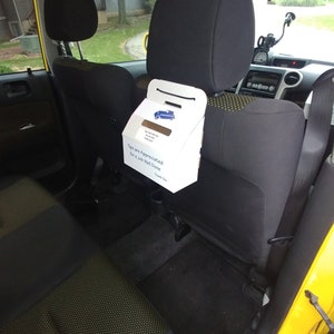 May include: A yellow car's backseat with a cardboard tip jar attached to the headrest. The tip jar has the text "Tips are Appreciated for a Job Well Done. Thank You."
