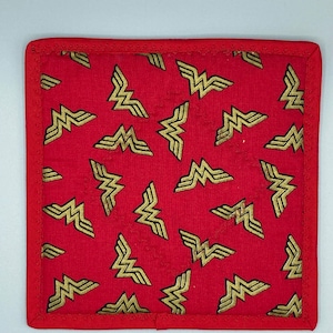 May include: A square, red Wonder Woman-themed pot holder with a repeating gold logo pattern. The edges are trimmed with a red border. The pot holder is likely made of fabric and is designed to protect surfaces from heat.