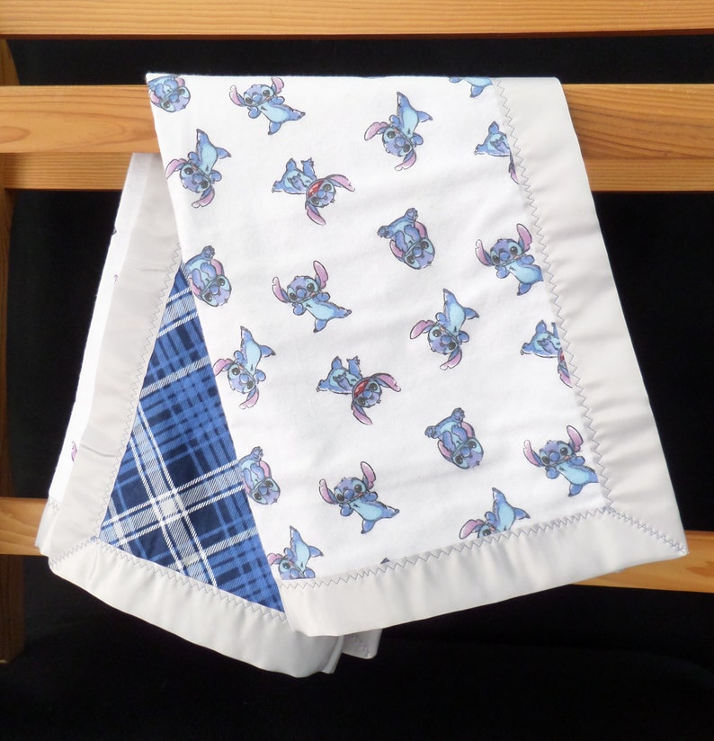 Puede incluir: Una manta para beb&eacute; blanca con un patr&oacute;n repetido de un personaje de dibujos animados azul. La manta tiene una esquina de cuadros azules y est&aacute; bordeada con un borde de sat&eacute;n blanco. La manta est&aacute; doblada sobre una estructura de madera.