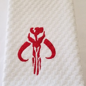 K&ouml;nnte beinhalten: Wei&szlig;es, strukturiertes K&uuml;chentuch mit einem rot gestickten Mandalorianer-Sch&auml;delsymbol. Das Handtuch hat ein Rautenmuster und ist gefaltet. Das rote Symbol ist auf dem Handtuch zentriert.