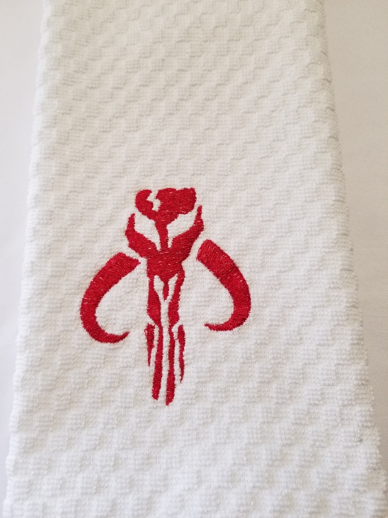 K&ouml;nnte beinhalten: Wei&szlig;es, strukturiertes K&uuml;chentuch mit einem rot gestickten Mandalorianer-Sch&auml;delsymbol. Das Handtuch hat ein Rautenmuster und ist gefaltet. Das rote Symbol ist auf dem Handtuch zentriert.