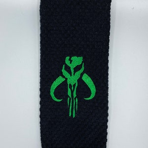 K&ouml;nnte beinhalten: Schwarzes Handtuch mit strukturiertem Gewebe. Ein gr&uuml;n gesticktes Design eines Sch&auml;dels mit gebogenen H&ouml;rnern ist auf dem Handtuch zentriert. Das Handtuch ist rechteckig und scheint aus einem weichen Material zu bestehen.