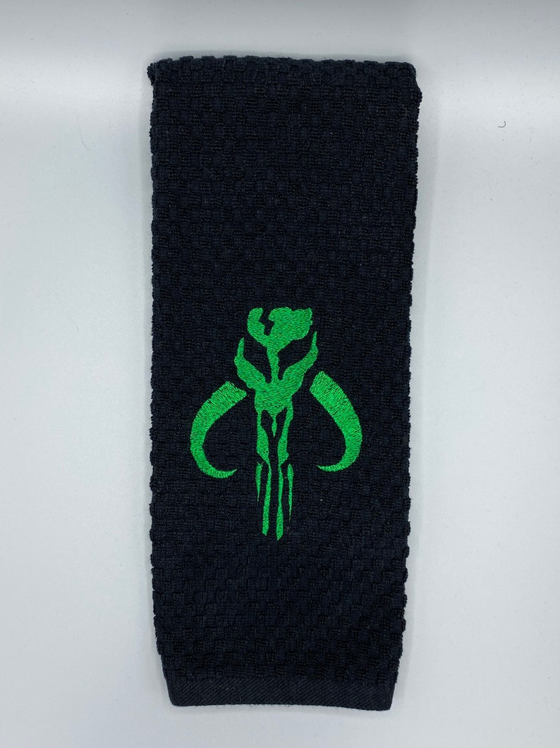 K&ouml;nnte beinhalten: Schwarzes Handtuch mit strukturiertem Gewebe. Ein gr&uuml;n gesticktes Design eines Sch&auml;dels mit gebogenen H&ouml;rnern ist auf dem Handtuch zentriert. Das Handtuch ist rechteckig und scheint aus einem weichen Material zu bestehen.