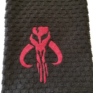 K&ouml;nnte beinhalten: Ein schwarzes Handtuch mit einem strukturierten Muster. Ein rotes gesticktes Emblem befindet sich auf dem Handtuch. Das Emblem ist ein stilisiertes Design mit gebogenen H&ouml;rnern und vertikalen Linien, das einem Sch&auml;del &auml;hnelt.