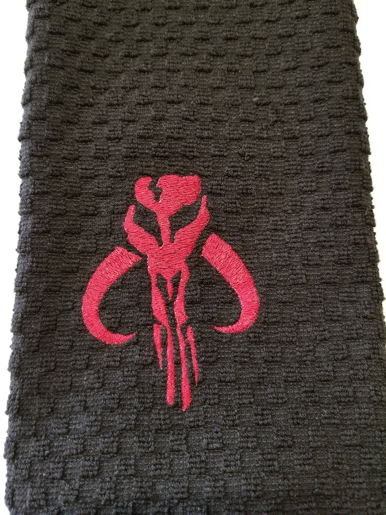 K&ouml;nnte beinhalten: Ein schwarzes Handtuch mit einem strukturierten Muster. Ein rotes gesticktes Emblem befindet sich auf dem Handtuch. Das Emblem ist ein stilisiertes Design mit gebogenen H&ouml;rnern und vertikalen Linien, das einem Sch&auml;del &auml;hnelt.