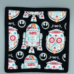 Può includere: Presina quadrata nera con un motivo ripetuto di figure di robot stilizzate e disegni di teschi di zucchero in bianco, rosso, arancione e blu. La presina ha un bordo nero.
