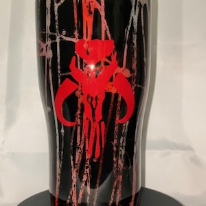 May include: A black insulated tumbler featuring a red Mandalorian skull emblem. The tumbler has a black and red splatter design. The lid is black with a silver rim. The cylindrical tumbler sits on a black base.