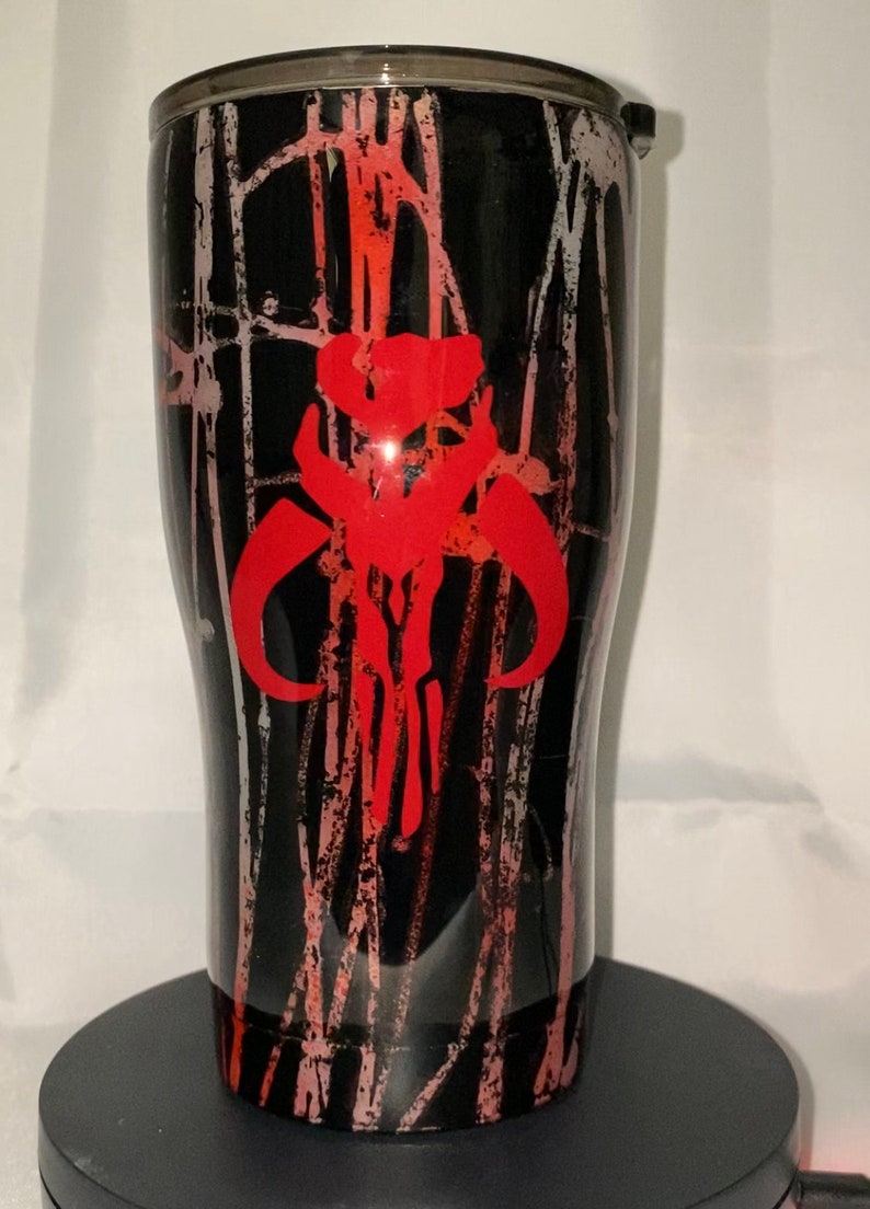May include: A black insulated tumbler featuring a red Mandalorian skull emblem. The tumbler has a black and red splatter design. The lid is black with a silver rim. The cylindrical tumbler sits on a black base.