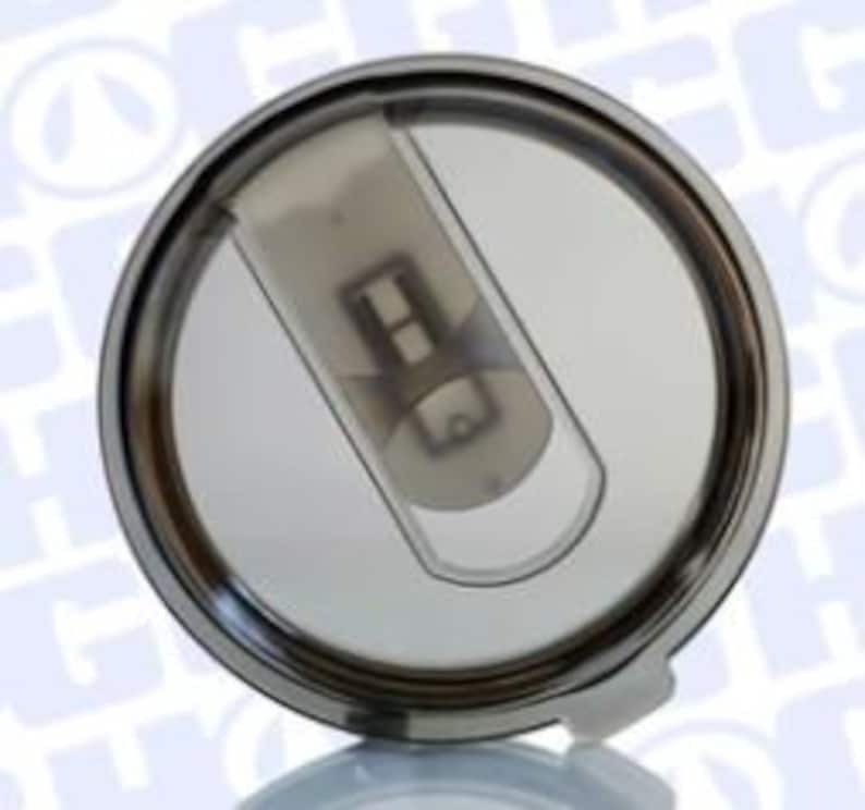 May include: A circular, stainless steel tumbler lid with a clear, sliding closure. The lid features a dark grey, translucent plastic slider and a small white tab. Designed to fit a tumbler and keep drinks secure.