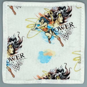 May include: Square white fabric featuring a Wonder Woman design. The pattern includes illustrations of Wonder Woman, the words "POWER" and "WONDER WOMAN", and the Wonder Woman logo. The edges are finished with a white border and a decorative stitch.