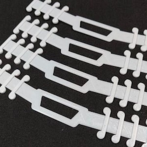 May include: Close-up of four white plastic mask extenders. Each extender has a series of small, rounded hooks on each end and a rectangular opening in the center. The extenders are arranged diagonally against a dark background.