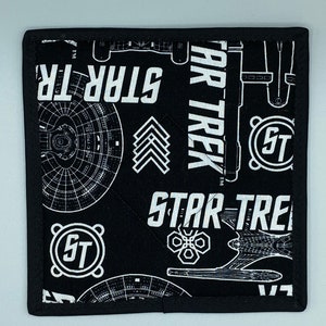 May include: Square black and white Star Trek themed pot holder. The design includes the words "STAR TREK" in large white letters, along with schematics of the starship Enterprise and other Star Trek symbols. The pot holder has a black border.