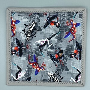 Puede incluir: Artículo de tela gris cuadrada con un patrón repetido de personajes de Spider-Man en varias poses. El diseño incluye las palabras "Spider-Man: Into the Spider-Verse". El borde es gris con un borde cosido.