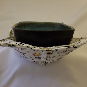 May include: A black square ceramic bowl with a teal interior sits inside a white fabric bowl cosy. The cosy is patterned with black and red cartoon illustrations and text. The cosy is designed to protect hands from hot bowls.